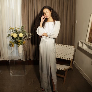 wearing a long powder blue dress a model stands facing the camera in a hotel room. She holds one hand up to her face showing the gathered cuff and the other arm is crossed over her body revealing the stretchy, gathered waistband and satin tie.