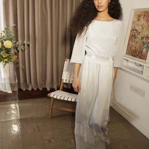 A lady with big curly hair walks towards the camera wearing a high neck, loose fitting dress. The long skirt flows as she walks and the sleeves fall to just over elbow length. The dress is pulled in at the waist with a gathered waistband.