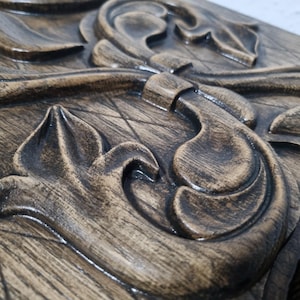 Può includere: Primo piano di un pannello decorativo in legno con un design intagliato dettagliato. Il pannello presenta forme organiche e motivi floreali a spirale in varie tonalità di marrone, con visibile venatura del legno. Il design è in rilievo.