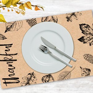 K&ouml;nnte beinhalten: Ein braunes Platzset mit einem schwarz-wei&szlig;en Blattdesign und dem Wort "Thankful" in Schwarz. Ein wei&szlig;er Teller, eine Gabel und ein Messer liegen auf dem Platzset.