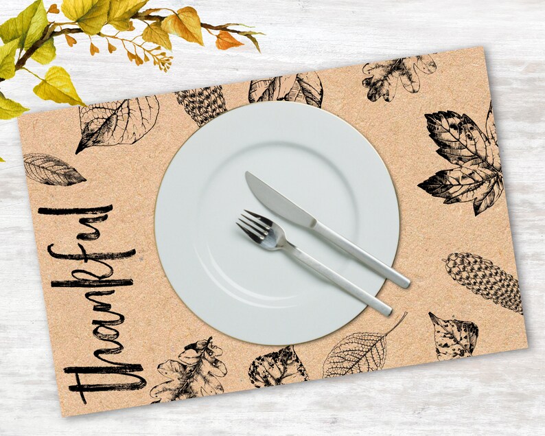 K&ouml;nnte beinhalten: Ein braunes Platzset mit einem schwarz-wei&szlig;en Blattdesign und dem Wort "Thankful" in Schwarz. Ein wei&szlig;er Teller, eine Gabel und ein Messer liegen auf dem Platzset.