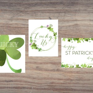 May include: Three St. Patrick's Day cards on a wooden surface. One card shows a green watercolor shamrock with the word "LUCKY". Another card has the words "Lucky Us" in a green floral wreath. The third card says "happy ST PATRICK'S day".