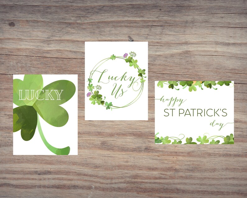 May include: Three St. Patrick's Day cards on a wooden surface. One card shows a green watercolor shamrock with the word "LUCKY". Another card has the words "Lucky Us" in a green floral wreath. The third card says "happy ST PATRICK'S day".