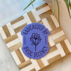 May include: A sticker with the text "RADICALIZED BY BASIC DECENCY" in white lettering on a light purple background. The sticker features a black floral design. The sticker is placed on a white and tan woven coaster.