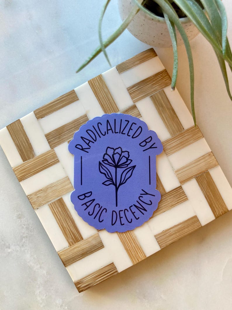 May include: A sticker with the text "RADICALIZED BY BASIC DECENCY" in white lettering on a light purple background. The sticker features a black floral design. The sticker is placed on a white and tan woven coaster.