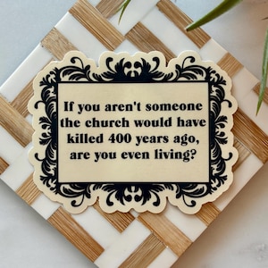 May include: A sticker with a black ornate border and text that reads, "If you aren't someone the church would have killed 400 years ago, are you even living?" The sticker is on a white and tan patterned surface.