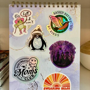 Op de afbeelding: Een spiraalgebonden notitieboek met een lichtpaarse en witte gemarmerde kaft, voorzien van zes stickers: een kikker met een roze cowboyhoed, een vogel, een pingu&iuml;n met zonnebril, drie heksen in een bos, een smeltende smiley en een zonsondergang.