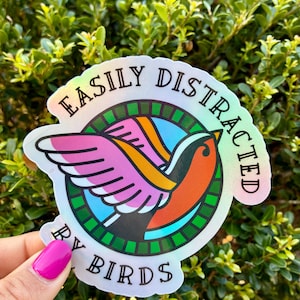 May include: Holographic sticker with a colorful bird design. The bird has pink wings, an orange body, and is set against a blue background. The sticker reads "EASILY DISTRACTED BY BIRDS" in a circular layout, perfect for bird lovers.