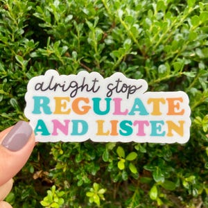 May include: A white sticker with the words "alright stop REGULATE AND LISTEN" in a colorful font. The sticker is held in front of a green leafy background.
