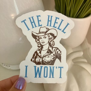 May include: A white sticker with a vintage illustration of a woman in a cowboy hat and jacket. The text "THE HELL I WON'T" is printed in blue, adding a bold statement to the design. The sticker is held by a hand with a purple nail.