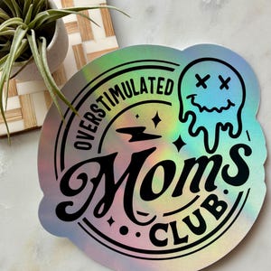 Op de afbeelding: Een ronde holografische sticker met de zwarte tekst OVERSTIMULATED MOMS CLUB. Het ontwerp bevat een smeltend smileygezicht met X-vormige ogen, een bliksemschicht en kleine sterren, allemaal gedrukt in een vet, donker lettertype op een reflecterend oppervlak.
