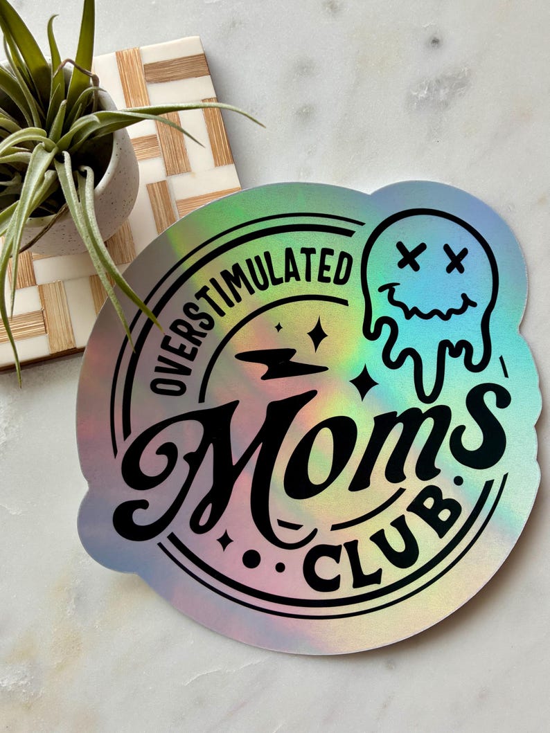Op de afbeelding: Een ronde holografische sticker met de zwarte tekst OVERSTIMULATED MOMS CLUB. Het ontwerp bevat een smeltend smileygezicht met X-vormige ogen, een bliksemschicht en kleine sterren, allemaal gedrukt in een vet, donker lettertype op een reflecterend oppervlak.
