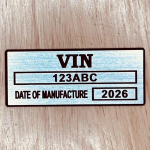 May include: A rectangular metal plate with the text "VIN", "123ABC", and "DATE OF MANUFACTURE 2026" in black. The background has a light blue brushed metal texture. The plate is on a wooden surface.