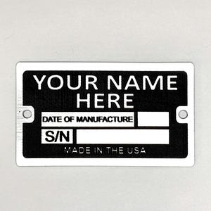May include: A rectangular black and white metal plate with the text "YOUR NAME HERE," "DATE OF MANUFACTURE," and "S/N." The bottom of the plate reads "MADE IN THE USA." The plate has two holes for mounting.