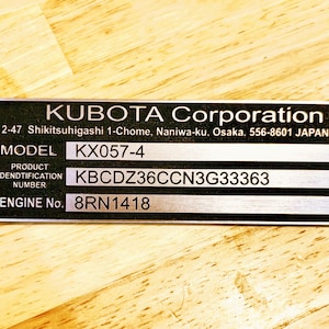 Puede incluir: Una placa metálica negra y plateada con el texto "KUBOTA Corporation" y la dirección en Japón. La placa incluye el número de modelo "KX057-4", el número de identificación del producto y el número de motor "8RN1418".
