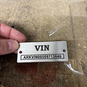 May include: A rectangular silver-colored metal identification plate with a brushed finish. The plate features the black printed text VIN at the top and the alphanumeric code ARKVIN00509713646 inside a black rectangular border below.