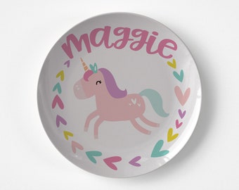May include: A white ceramic plate with a pink unicorn design and the name "Maggie" written in pink cursive. The unicorn has a purple mane and tail, a gold horn, and a pink body. The plate is decorated with a border of pink, yellow, and blue hearts.