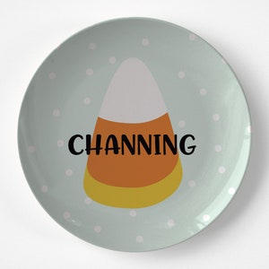 May include: A round, light blue plate with white polka dots. The plate features a candy corn graphic with white, orange, and yellow layers. The name "CHANNING" is printed in black across the candy corn.