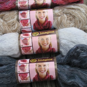 Könnte beinhalten: Vier Knäuel Imagine-Garn in verschiedenen Farben, darunter Rot, Beige, Silber und Blau. Jedes Knäuel hat ein Etikett mit einer Frau, die einen Schal trägt, und dem Wort "Imagine".