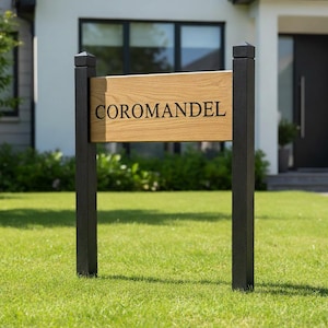 Può includere: Un cartello in legno con la scritta "COROMANDEL" in nero. Il cartello è realizzato in legno chiaro ed è sostenuto da due pali neri. Il cartello è posto in un giardino erboso.