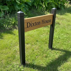 May include: A wooden sign with the text "Dixon House" in black lettering. The sign is mounted on two black posts with a pointed top, set in a grassy lawn. The sign is made of light brown wood.