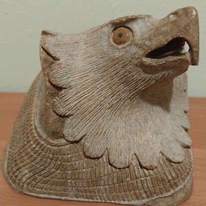 Könnte beinhalten: Eine geschnitzte Steinskulptur eines Adlerkopfes mit detaillierter Federstruktur. Das Auge des Adlers ist aus einem anderen Material gefertigt und hat eine hellbraune Farbe.