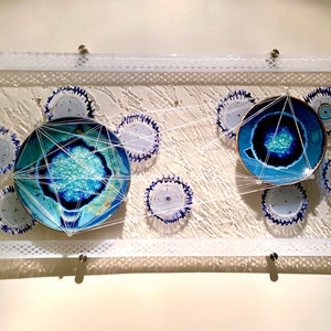 Può includere: Un pannello di vetro rettangolare bianco con due piatti in ceramica blu e bianca collegati da una rete di corde bianche. I piatti sono circondati da cerchi di ceramica blu e bianca più piccoli.