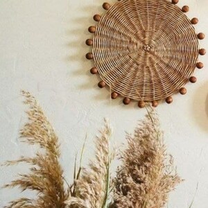 May include: A round woven wall decoration with a natural brown colour and a beaded border. The beads are a darker brown colour. The decoration is hanging on a white wall with dried pampas grass in the foreground.