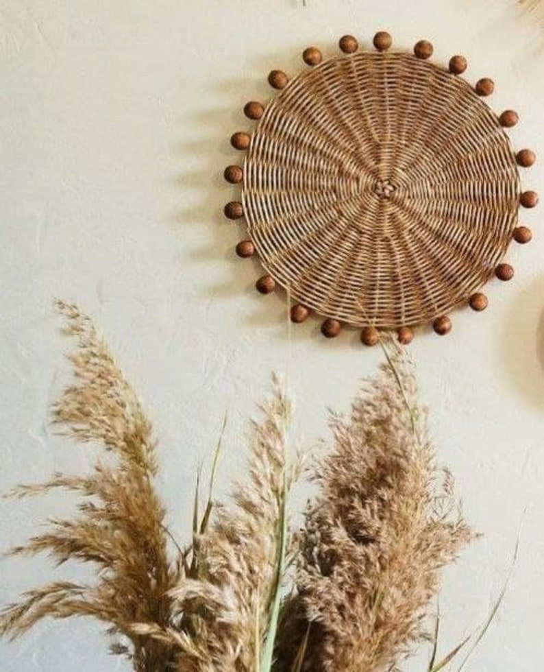 May include: A round woven wall decoration with a natural brown colour and a beaded border. The beads are a darker brown colour. The decoration is hanging on a white wall with dried pampas grass in the foreground.