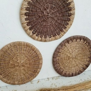 May include: Three round woven wall decor pieces in various sizes. The largest piece is brown with a natural woven border. The two smaller pieces are natural woven with a brown border.