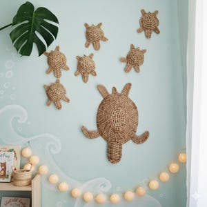 May include: Woven rattan turtle wall decorations in various sizes, mounted on a light blue wall with a wave design. The turtles are in shades of brown and tan. A string of warm-toned, round lights adds a soft glow to the scene.