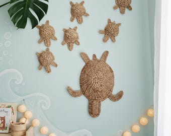 Puede incluir: Un conjunto de siete decoraciones de pared con forma de tortuga marina tejidas con fibras naturales de color tostado. El set incluye una tortuga grande y seis más pequeñas, con patrones de tejido circulares y aletas detalladas.