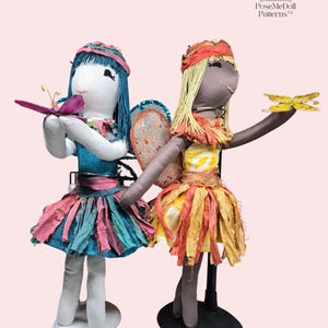 May include: Two handmade fabric dolls with wings, one with blue hair and a turquoise dress, the other with blonde hair and a yellow dress. Both dolls are holding a butterfly in their hands. The dolls are standing on a white and black stand. PoseMeDoll Patterns.