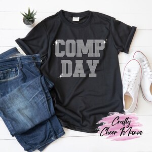 May include: Black t-shirt with the text "COMP DAY" in a rhinestone design. The shirt is paired with blue jeans and white sneakers. A small potted plant is visible in the background.