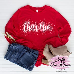 Może przedstawiać: Czerwona koszula z długim rękawem z napisem "Cheer Mom" w kolorze białym z białymi akcentami brokatowymi. Koszula leży na białym tle z niebieskimi dżinsami i brązowymi butami.