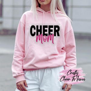 以下が含まれることがあります： 黒とピンクの「CHEER MOM」の文字がラインストーンで装飾された、明るいピンク色のパーカー。パーカーにはフロントポケットとドローストリング付きのフードが付いています。「Crafty Cheer Mama」の文字が右下にあります。