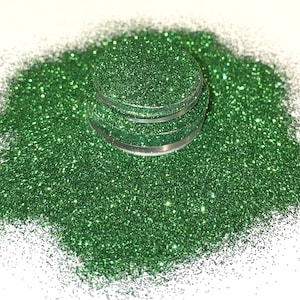 May include: A close-up of a jar of green glitter. The glitter is loose and has a fine texture. The jar is clear glass and has a silver lid.