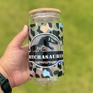 May include: A clear glass can-shaped tumbler with a bamboo lid. The tumbler features a camouflage pattern with a dinosaur graphic and the text "BITCHASAURUS" in a black banner.