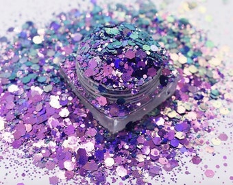 May include: A clear, square container filled with iridescent glitter in shades of purple, blue, and teal. The glitter is scattered around the container, creating a sparkling effect. The glitter is in various shapes and sizes, reflecting light.