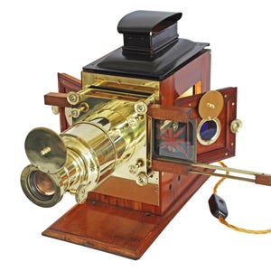 Puede incluir: Proyector de linterna mágica antiguo con lente de latón y carcasa de madera. El proyector tiene una parte superior negra, puertas de madera y una base. La lente es grande y prominente, y el proyector tiene una estética vintage.