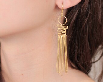 Può includere: Orecchini color oro dal design unico. Gli orecchini presentano un elemento superiore circolare, una sezione centrale a spirale e molteplici pendenti verticali a forma di barra. La persona che indossa gli orecchini ha i capelli castani.