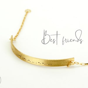 Könnte beinhalten: Goldfarbene Halskette mit gebogenem Barrenanhänger. Der Anhänger ist mit Morsecode versehen, und die Worte "Best friends" sind kursiv geschrieben. Die Halskette hat eine zarte Kette.