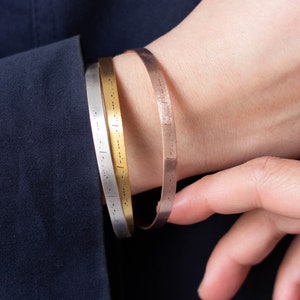 Op de afbeelding: Drie metalen manchetarmbanden in zilver, goud en roségoud. Elke armband heeft een reeks kleine, gelijkmatig verdeelde markeringen. De armbanden worden om een pols gedragen, met een donkerblauw kledingstuk op de achtergrond.