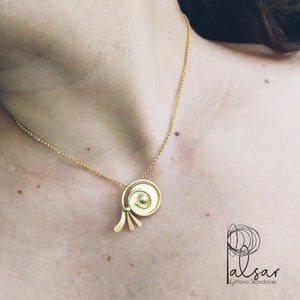 Op de afbeelding: Een gouden ketting met een unieke hanger met een cirkelvormig ontwerp met een kleinere cirkel in het midden. De hanger heeft een gebogen vorm en drie kleine, dunne, gebogen stukken die naar beneden uitstrekken.