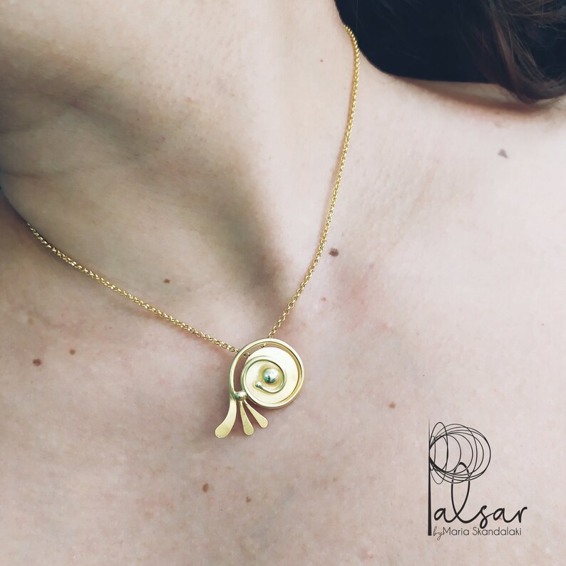 Op de afbeelding: Een gouden ketting met een unieke hanger met een cirkelvormig ontwerp met een kleinere cirkel in het midden. De hanger heeft een gebogen vorm en drie kleine, dunne, gebogen stukken die naar beneden uitstrekken.