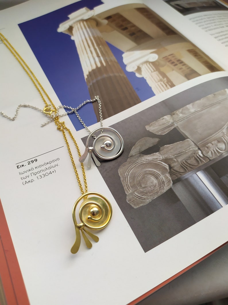 Op de afbeelding: Twee gouden en zilveren kettingen met ronde hangers. De hangers hebben een spiraalvormig ontwerp en een kleine bol in het midden. De kettingen worden getoond op een boek met een afbeelding van een kolom.