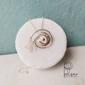 Op de afbeelding: Zilveren ketting met een spiraalvormige hanger met een getextureerd oppervlak en een kleine cirkel in het midden. De ketting ligt op een wit marmeren oppervlak.