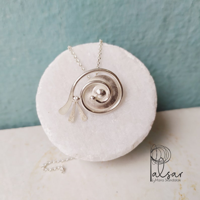 Op de afbeelding: Zilveren ketting met een spiraalvormige hanger met een getextureerd oppervlak en een kleine cirkel in het midden. De ketting ligt op een wit marmeren oppervlak.