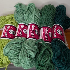 May include: Five skeins of Aunt Lydia's yarn arranged in a gradient from light lime green to deep forest green. Each skein features a white and red paper label with the text Aunt Lydia's, American Thread Co., and 70 yards, showcasing various green hues.