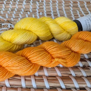 May include: Two skeins of fingering weight yarn, one bright yellow and one vibrant orange, each featuring a white paper label with black text indicating 20 grams and 92 yards. The yarn is twisted into a soft, textured coil, showcasing the fiber.